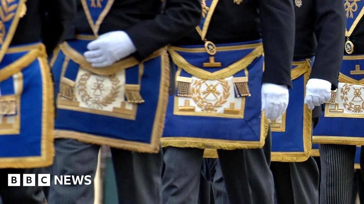 Masons march in a procession. Pictured are men from waist height wearing white gloves and wearing the Freemasons coat of arms embraided with gold thread on a royal blue background
