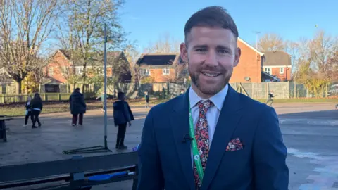 Headteacher James Cassidy is wearing a blue suit, flowery tie and smiling at the camera. He is standing in the playground and there are children playing behind him.