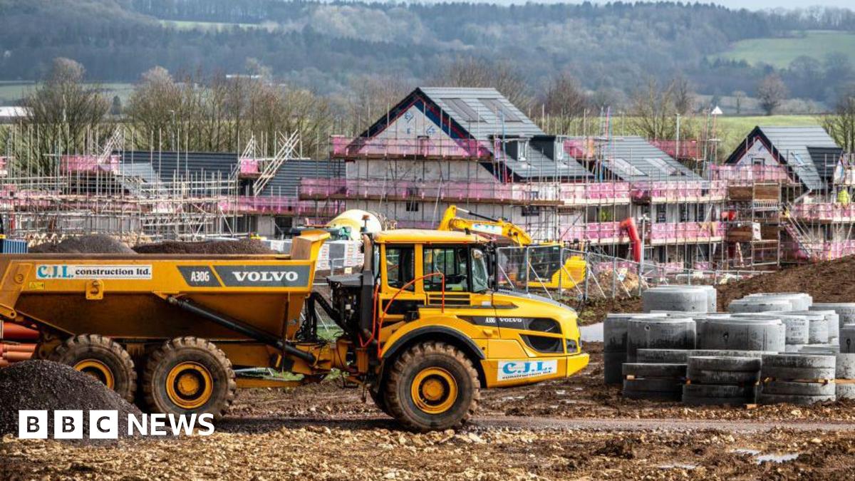 Four new detached houses surrounded by scaffolding rise from the ground. In the distance there are forested green slopes. A yellow digger is in the foreground.