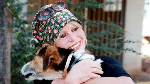 Charly Hel/Prestige/Getty Images Brigitte Bardot, wearing a head scarf and sunglasses on top of her head, smiles as she hugs a dog 