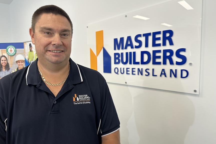 A dark-haired man in a branded polo shirt stands in an office with a sign that says "Master Builders Queensland" on the wall.