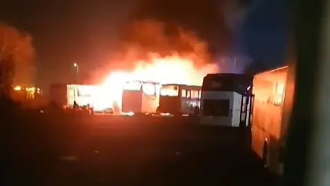 Inverclyde Community and Road Watch Large fire engulfing several buses in a dark outdoor lot at night, with thick black smoke rising into the sky.