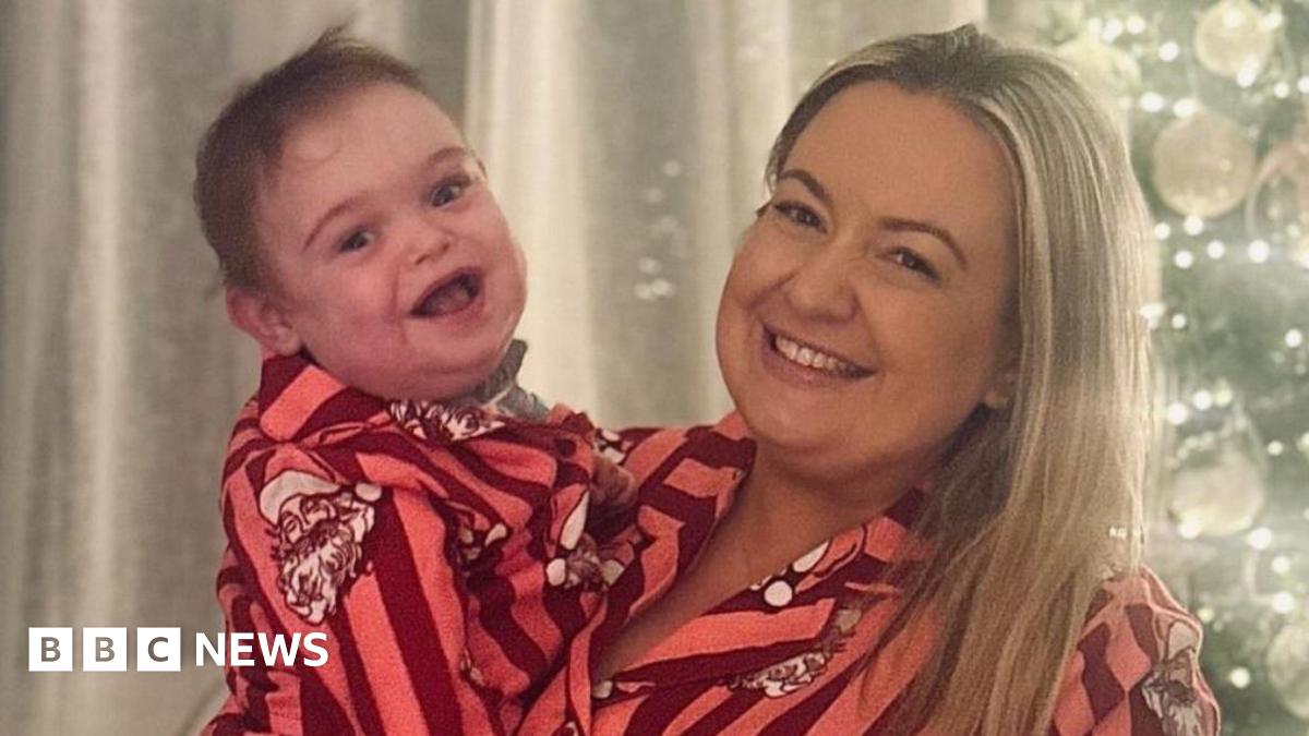 A blonde woman wearing pyjamas is holding her baby daughter. They are standing in front of a Christmas.