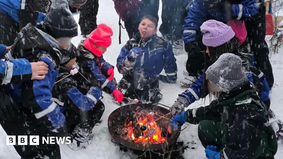 A group of adults and children sitting around a camp fire in the snow, roasting marshmallows. The children's faces are obscured so they cannot be identified.