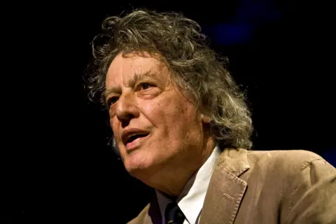 Getty Images Sir Tom Stoppard has wavy, shoulder-length hair and is wearing a light brown corduroy jacket over a white shirt and a dark tie. The background is dark, suggesting a stage or event setting with focused lighting on Sir Tom. 