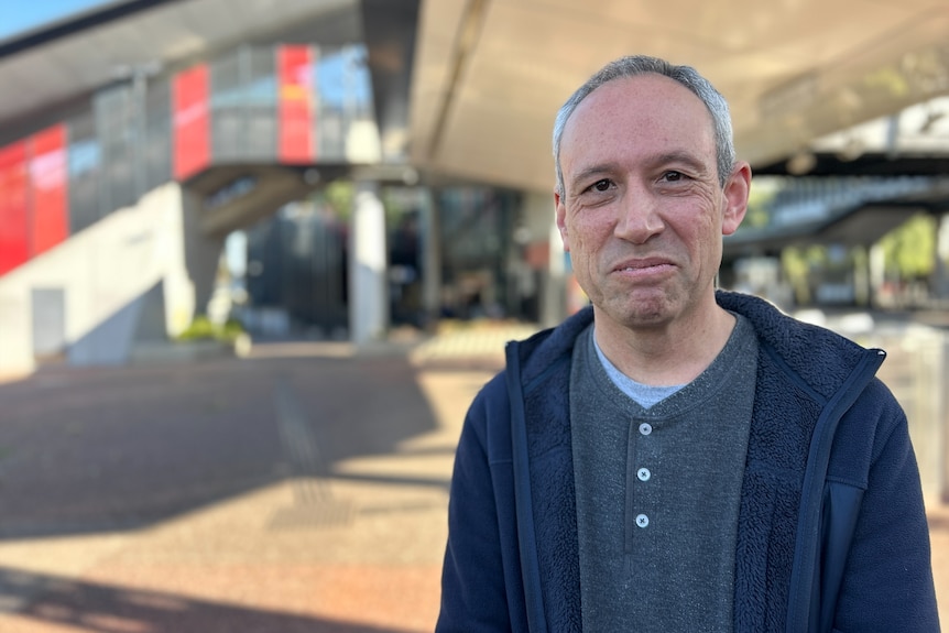 A man with grey hair in a grey shirt with dark jumper stares at the camera with a closed lip smile.