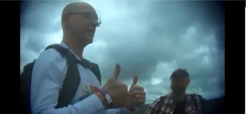 BBC Mr Lawler, a bald man wearing a white long-sleeve t-shirt with a partially-visible graphic on the front, gives a double thumbs-up gesture towards somebody not in frame. He is wearing a rucksack and standing next to another man, whose face is blurred.