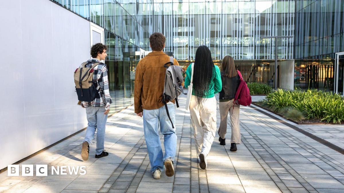 Four people with backpacks walk along a paved path towards a modern glass building. The group of students is seen from behind, wearing casual clothes, and the setting includes greenery and reflective windows.