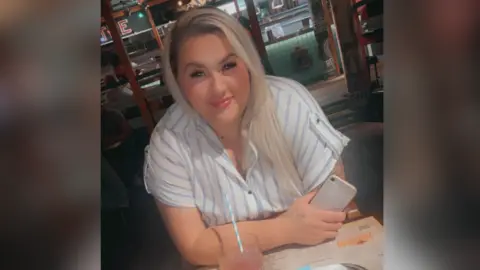 Supplied A close up of Sophie sitting at a table with a drink in front of her. It is in a glass with a blue and white straw. Sophie holds a phone in her hand and wears a short sleeved blouse with blue stripes. She has long hair that is blonde and dark.