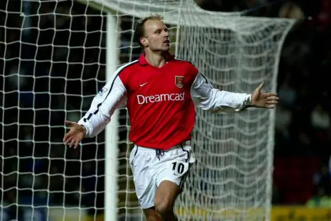 PA Media Dennis Bergkamp in a red Arsenal shirt celebrates a goal. His arms are outstretched in front of the net