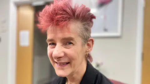 Mark Norman/BBC Lucinda Wentworth smiling at the camera wearing a black blouse with her hair dyed red 