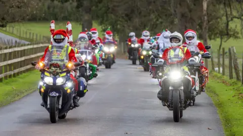 Del Hickey A group of people all dressed as Santa ride together on motorbikes down a road as part of the charity event. All the bikes are also decorated with tinsel. 