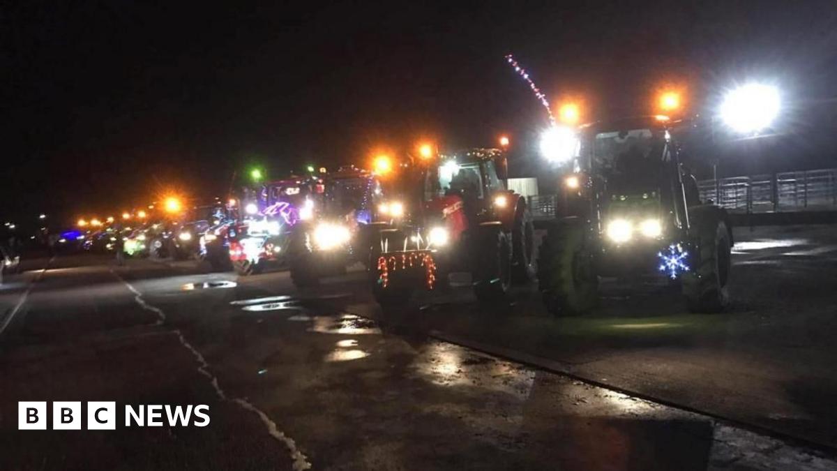 Several tractors are parked alongside one another, in the dark, on a large tarmac area. They have headlights on full and multiple Christmas lights as decoration.
