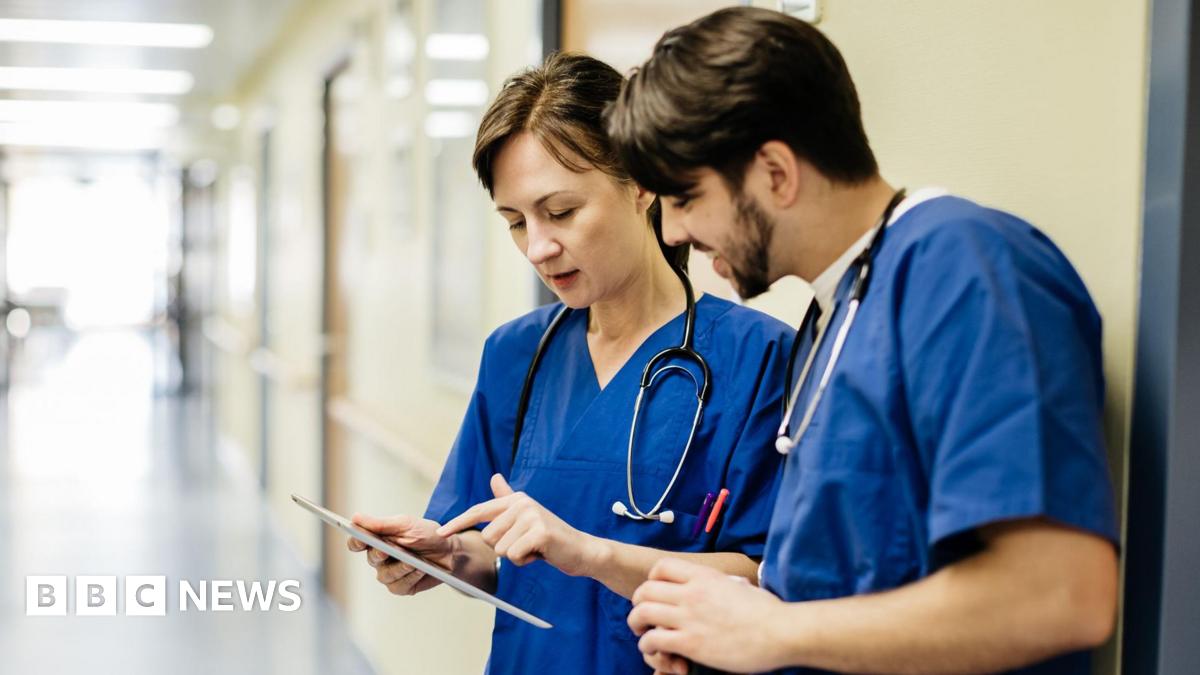 A male and female doctor, both wearing blue scrubs, standing in an empty hospital hallway examining an iPad