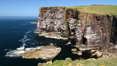 Getty Images Sandstone sea cliffs rise from the dark blue waters of the sea.