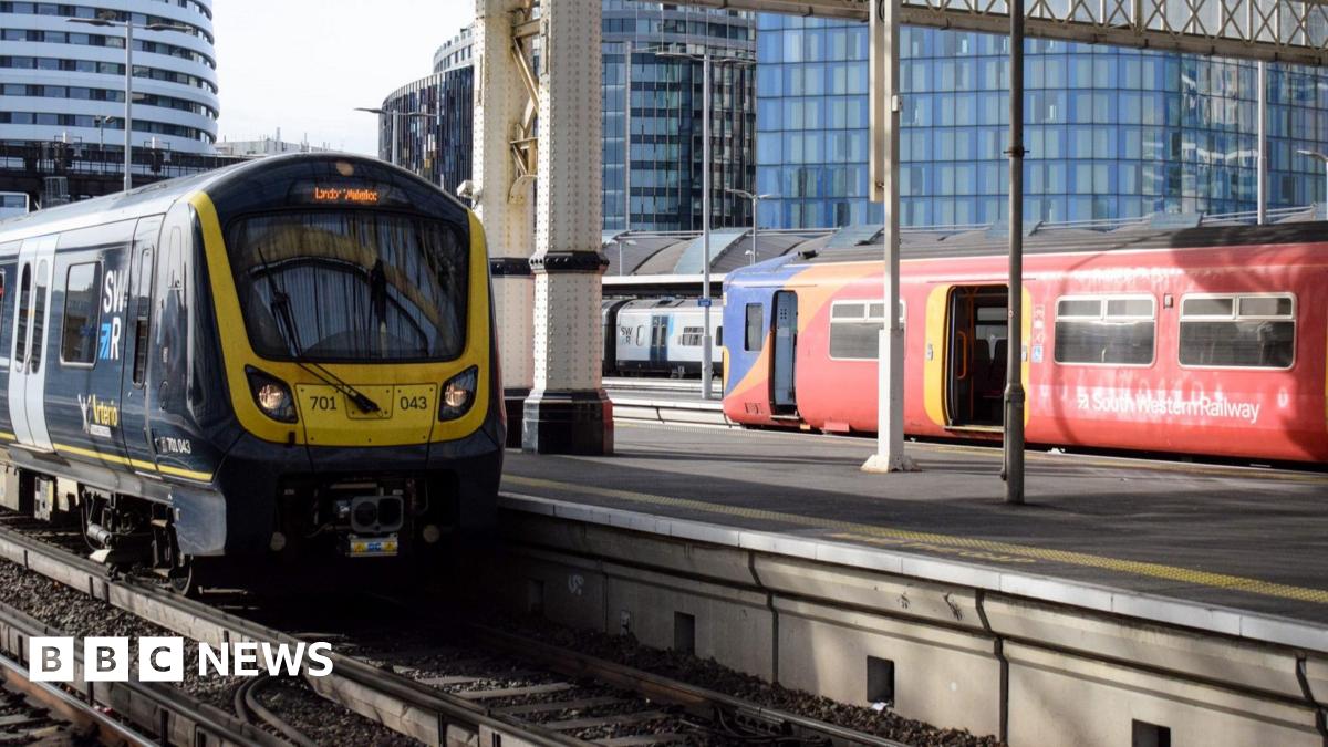 A red and yellow train with South West Railway branding at a station with another behind it