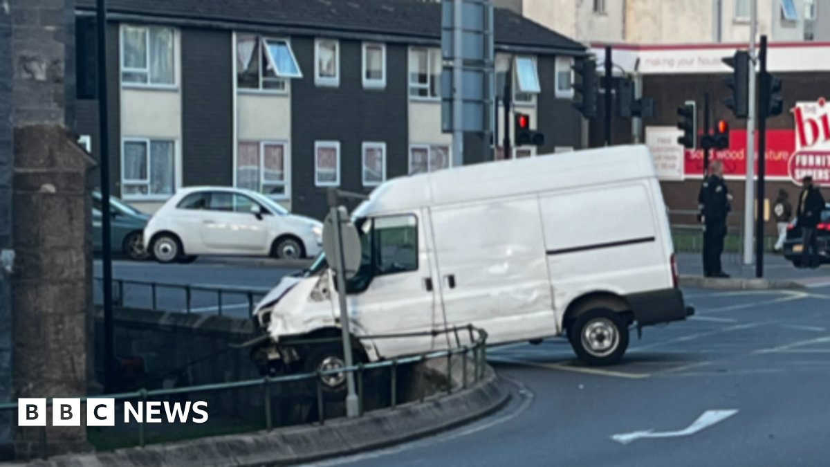 A derelict church on a busy roundabout in a city centre. Cars are travelling on the road and roundabout around the church. It is located in front of a large shopping mall. Green traffic lights are on the road around the church.
