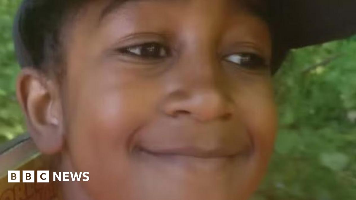 A photo of Adam where he is smiling. He has dark hair and brown eyes. Grass is visible in the background.