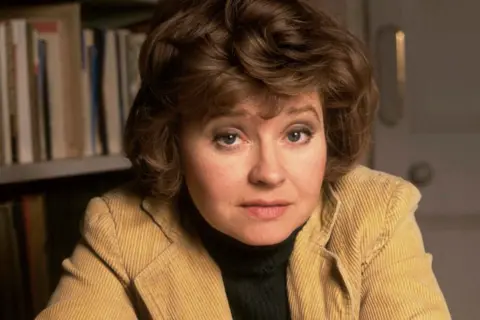 Getty Images Prunella Scales is wearing a light brown corduroy jacket over a dark turtleneck sweater is seated indoors. Behind her is a bookshelf filled with books, and to the right, part of a light-coloured door with a round handle is visible. The setting appears to be a cozy, study-like environment with warm lighting.
