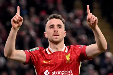 Getty Images Diogo Jota, wearing a red Liverpool FC jersey with yellow pinstripes and the club crest, is raising both arms with index fingers pointing upward in a celebratory gesture. The shirt features the sponsor logo “standard chartered” and a white collar detail. The background shows a blurred crowd in a stadium.
