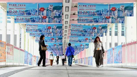 VCG via Getty Images Hongkongers walk on an overhead bridge festooned with blue posters advertising the 2025 Hong Kong Legco election campaign