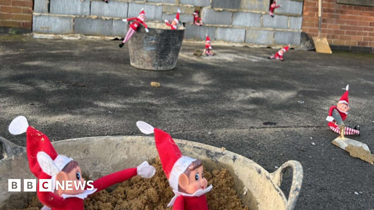 Paul Heeley is standing outside his office with his arms crossed and a mock-angry look on his face. Small colourful balls are piled up in the room. Red and white toy elves are sitting on the window sill.