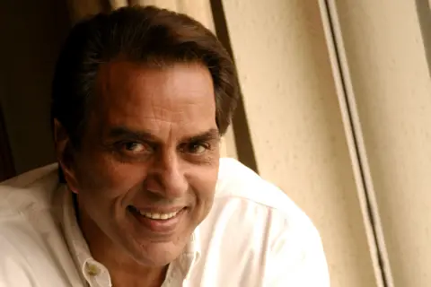 Getty Images Dharmendra wears a white button-up shirt, sitting indoors near a window.