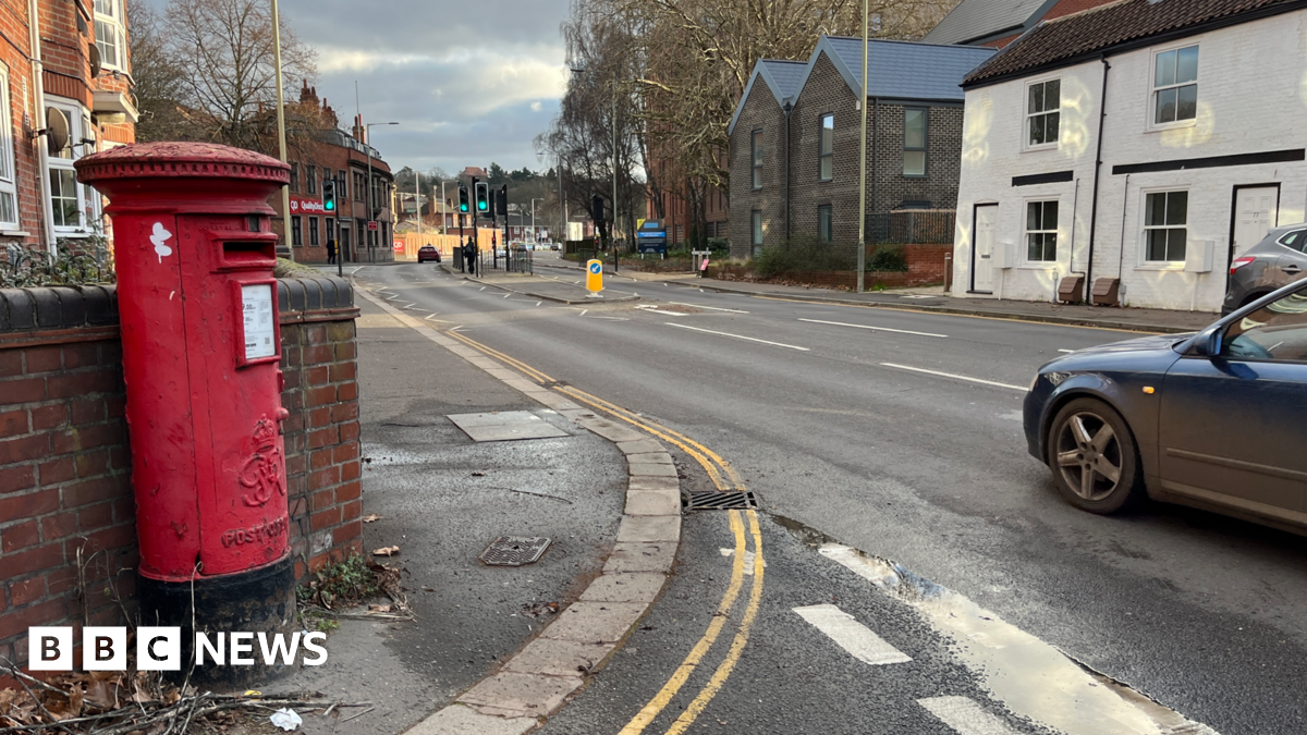 A red postbox is seen to the left of a suburban single-carriageway road, which is lined by houses and some trees, with traffic lights and a pedestrian crossing in the centre.