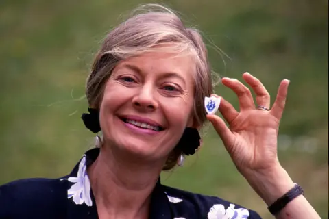 Biddy Baxter is holding up a small white Blue Peter badge featuring a blue shield emblem with a stylized design. She is wearing a dark garment with a floral pattern and a wristwatch on the left wrist. Behind her is an outdoor grassy area, suggesting the photo was taken in a natural setting.
