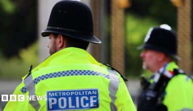 Two police officers in high vis face away from the camera and they have "Metropolitan Police" printed on their backs. One man has his back to the camera and another looks to the left. Both wear helmets.
