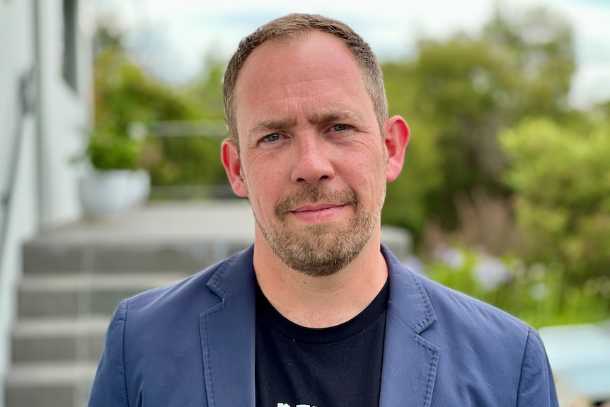 A white man with facial hair wearing a black tshirt and a dark blue blazer.