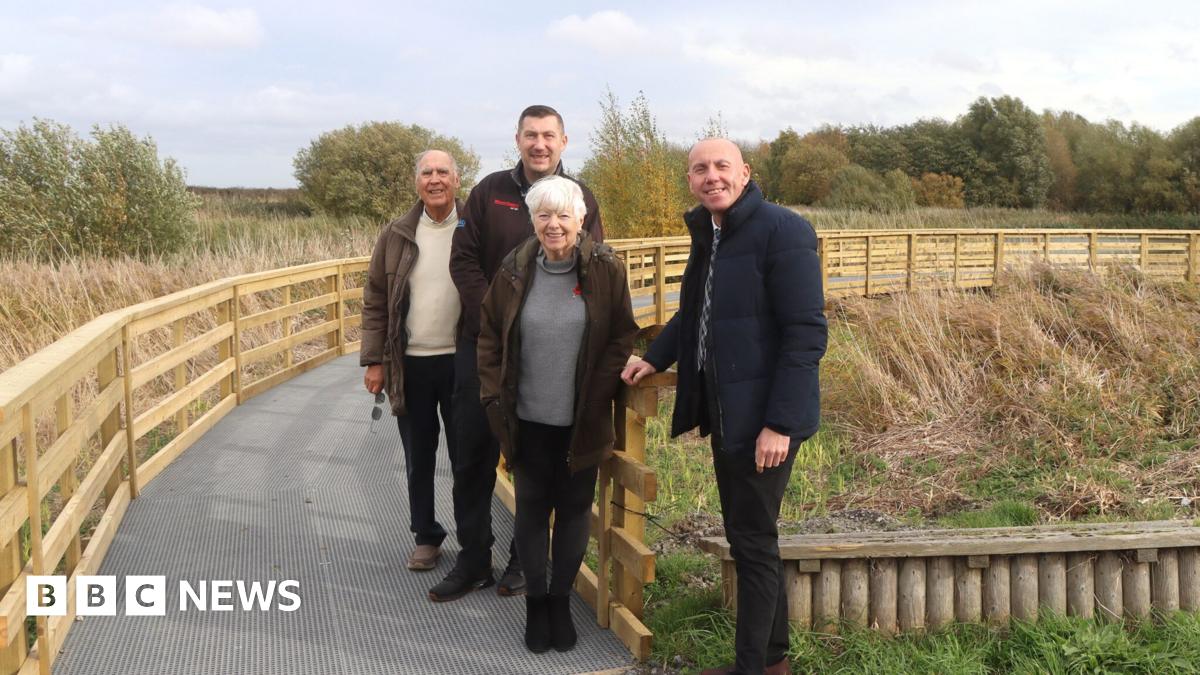 Three men and a woman pictured standing on a fenced boardwalk with trees in the background. They are all looking towards the camera and smiling.