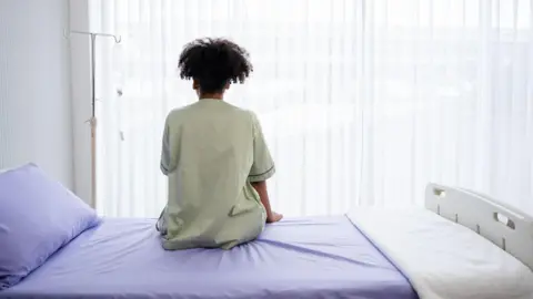 Getty Images via 24/7Studio A person in a pale green hospital gown sits on a hsopital bed, made up with a lilac bedspread, pillow case and white blanket. They are facing away from the camera towards the window, with a gurney left of them. 