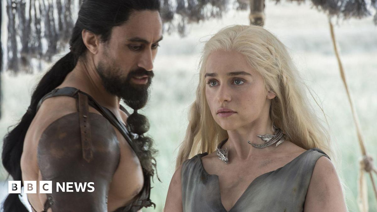 Joe Naufahu and Emilia Clarke, characters from Game of Thrones, dressed in fantasy medieval leather clothing. They are scantily clad but there appears to be snow on the ground behind them. Their skin is dirty.