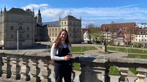 Claire Thomson Claire wearing a white long sleeved t-shirt and black dungarees standing looking over a town square with different styles of traditional German buildings in the background.