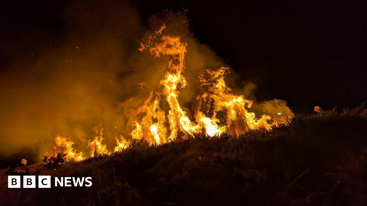An image of tall, bright orange and black flames coming off the moorland. It is dark and the bright flames contrast against the night sky. There is also a lot of smoke coming from the area. There is a firefighter to the left of the image.