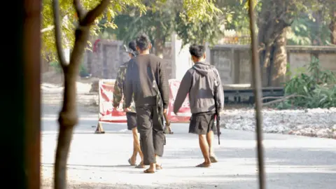 Lulu Luo/ BBC Two boys are carrying guns and walking down the street, their backs to the camera. 
