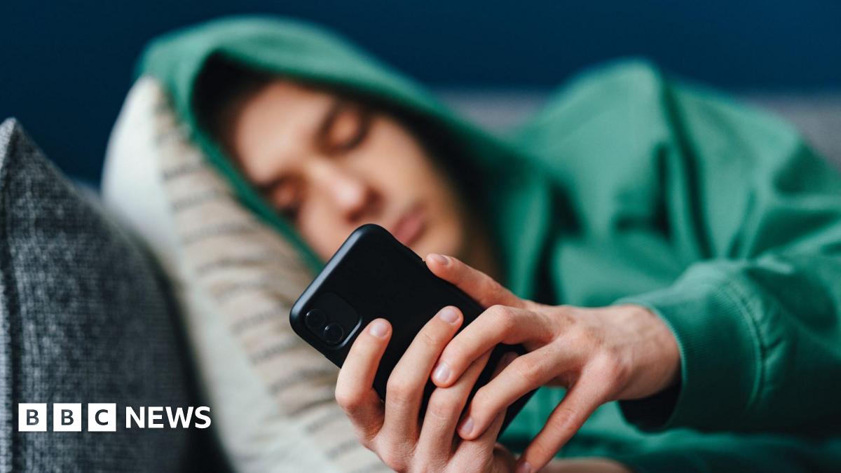 A teenager wearing a green hoody with the hood pulled up, lies on cushions on a sofa looking at his mobile phone
