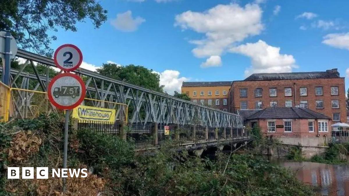 A steel footbridge over a river with some historic, brown-brick buildings in the background