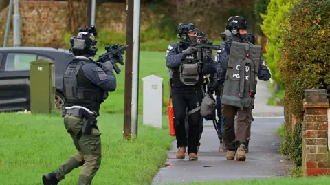 Dan Jessup Men in police tactical gear on a footpath surrounded by grass. Their mouths and noses are covered by balaclavas. One is seen carrying a rifle and another carries a shield. Another police officer with a rifle is stood on the grass.