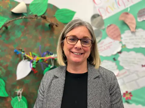 Curriculum manager Anne Clark wearing a pair of glasses, a grey blazer and a  black top. She is smiling and standing in front of a colourful nursery artwork wall. 