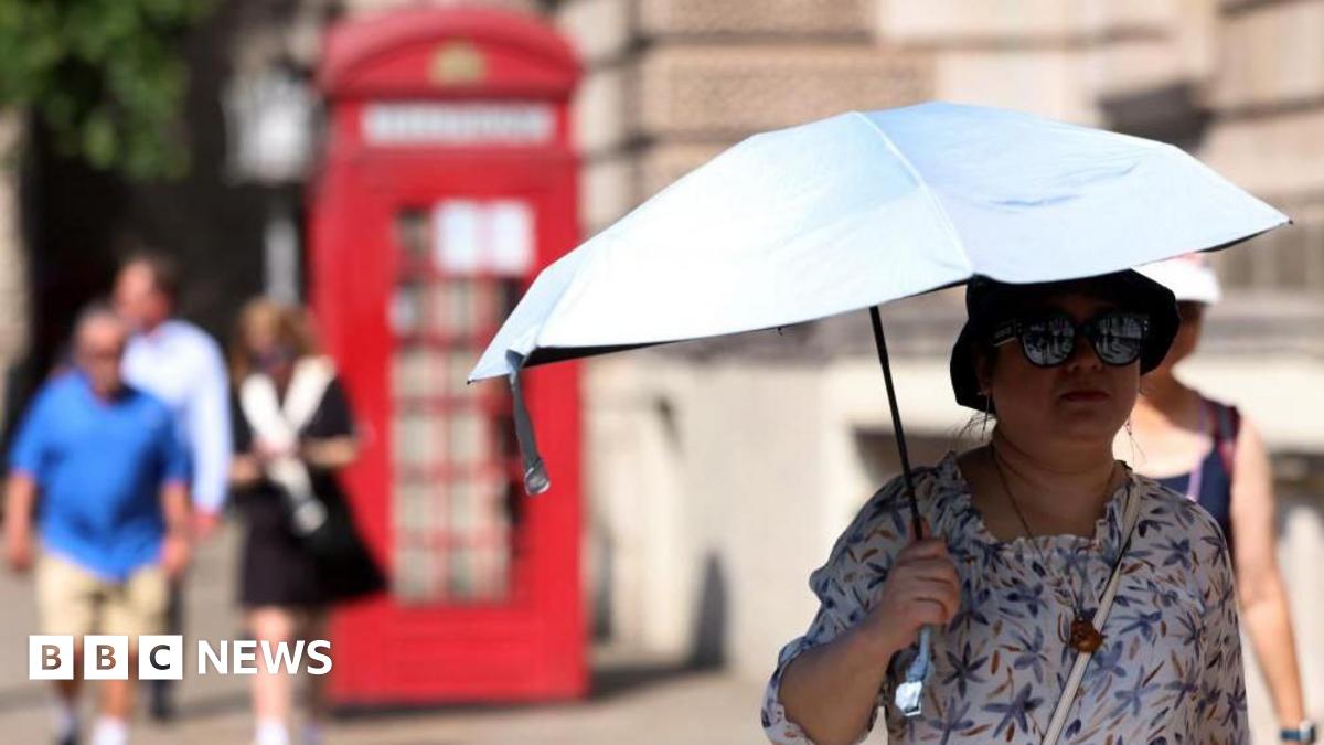 A woman, wearing a sunhat and sunglasses, holds an umbrella to shade herself as she walks along a street in London. There is a red telephone box behind her as well as a few other pedestrians.