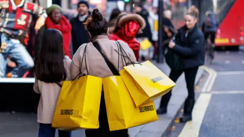EPA A woman walks along Oxford Street in central London with three bright yellow Selfridges shopping bags on her shoulders.