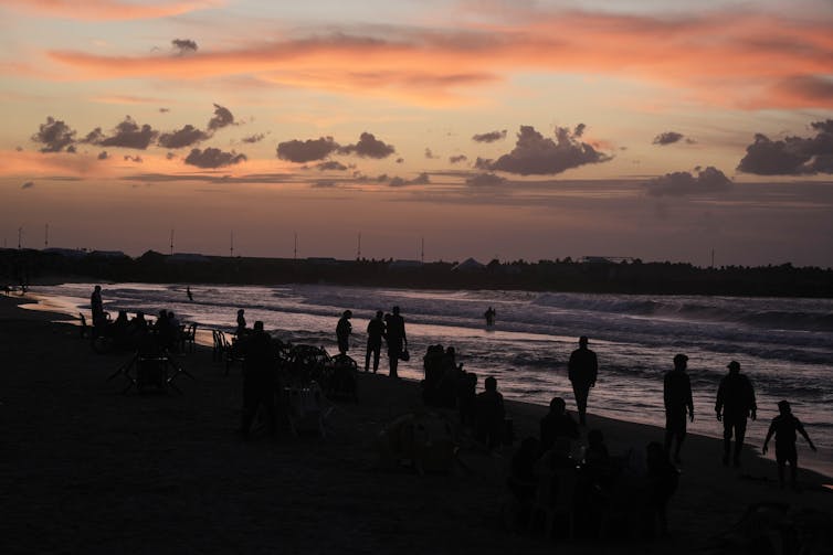 People seen walking at a beach against a purplish sky.