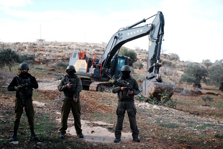 Three soldiers stand in front of a mechanical digger