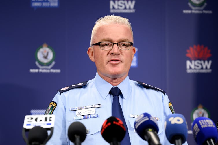 A man in a police shirt and tie speaks at a podium behind a row of microphones