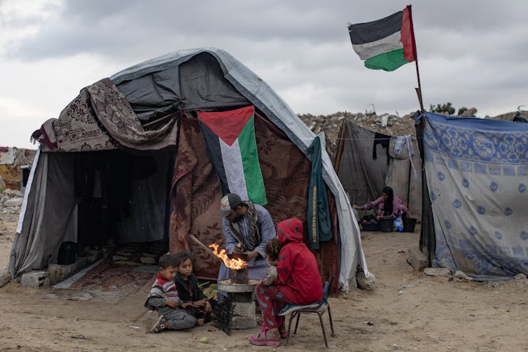 Palestinians dressed in warm clothing outside a makeshift shelter with a Palestinian flag in the background.