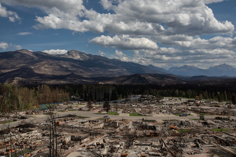 an aerial photo of destroyed buildings and rubble, mountains under a blue sky with white clouds in the background.