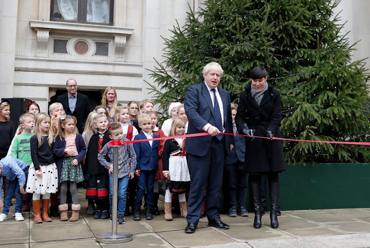 A man and women cut a red ribbon in front of a large tree.