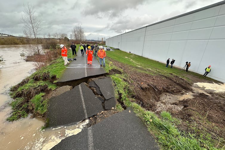 A paved bicycling path atop a levee is broken and slabs of asphalt pavement are tilted into a breach where water poured through.